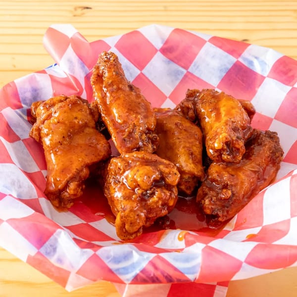 Six glazed chicken wings in a red and white checkered paper-lined basket on a wooden table.
