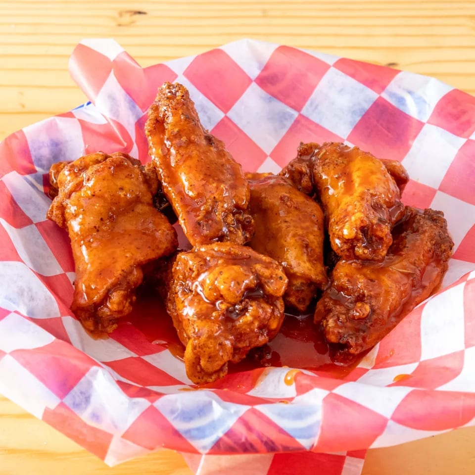 Six glazed chicken wings in a red and white checkered paper-lined basket on a wooden table.