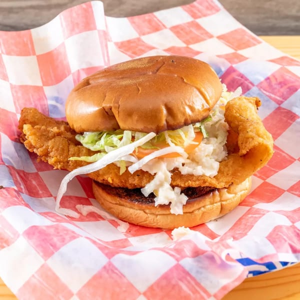 Crispy fried fish sandwich with coleslaw and lettuce on a bun, wrapped in red and white checkered paper.