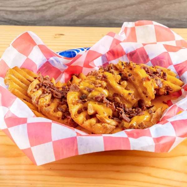 Waffle fries topped with melted cheese and bacon bits in a red and white checkered paper basket.