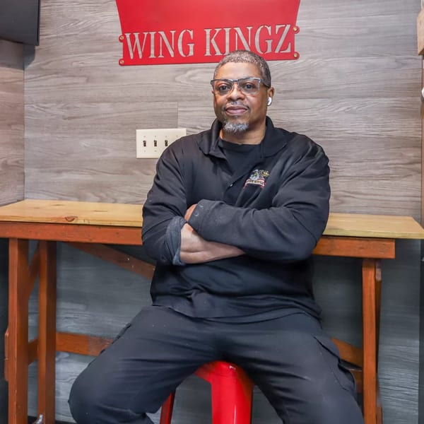 Man in black shirt and glasses sitting on a red stool under a "Wing Kingz" sign.