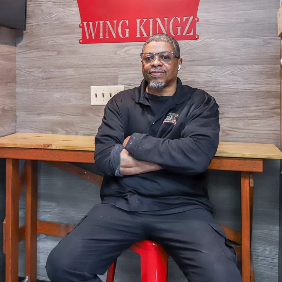 Man in black shirt and glasses sitting on a red stool under a "Wing Kingz" sign.