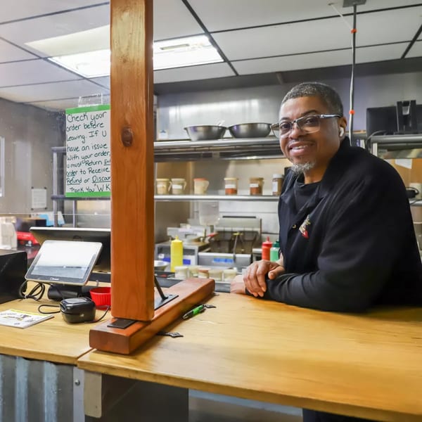 A smiling man in glasses and a black chef's coat leans on a wooden counter in a restaurant kitchen.